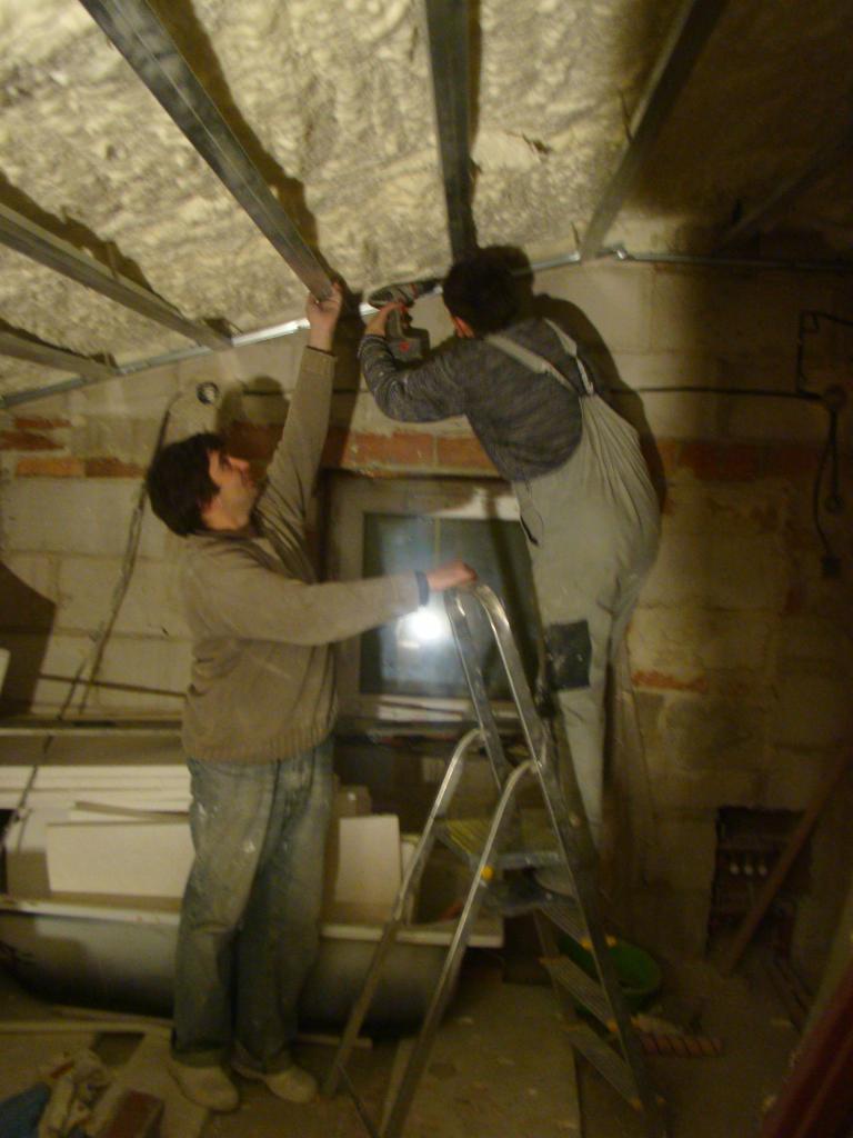 Montaż sufitu podwieszanego: Dwóch mężczyzn mocuje metalowe profile do ocieplonego stropu za pomocą wkrętarki, stojąc na aluminiowej drabinie w trakcie prac wykończeniowych.