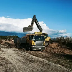 Żółta ciężarówka Scania i koparka Volvo na placu budowy, wykop pod instalacje widoczny w tle, błękitne niebo z chmurami.