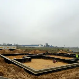 Wylany fundament pod budynek z hydroizolacją, ubijarka wibracyjna na piasku, wywrotka z piaskiem w tle, pochmurny dzień.