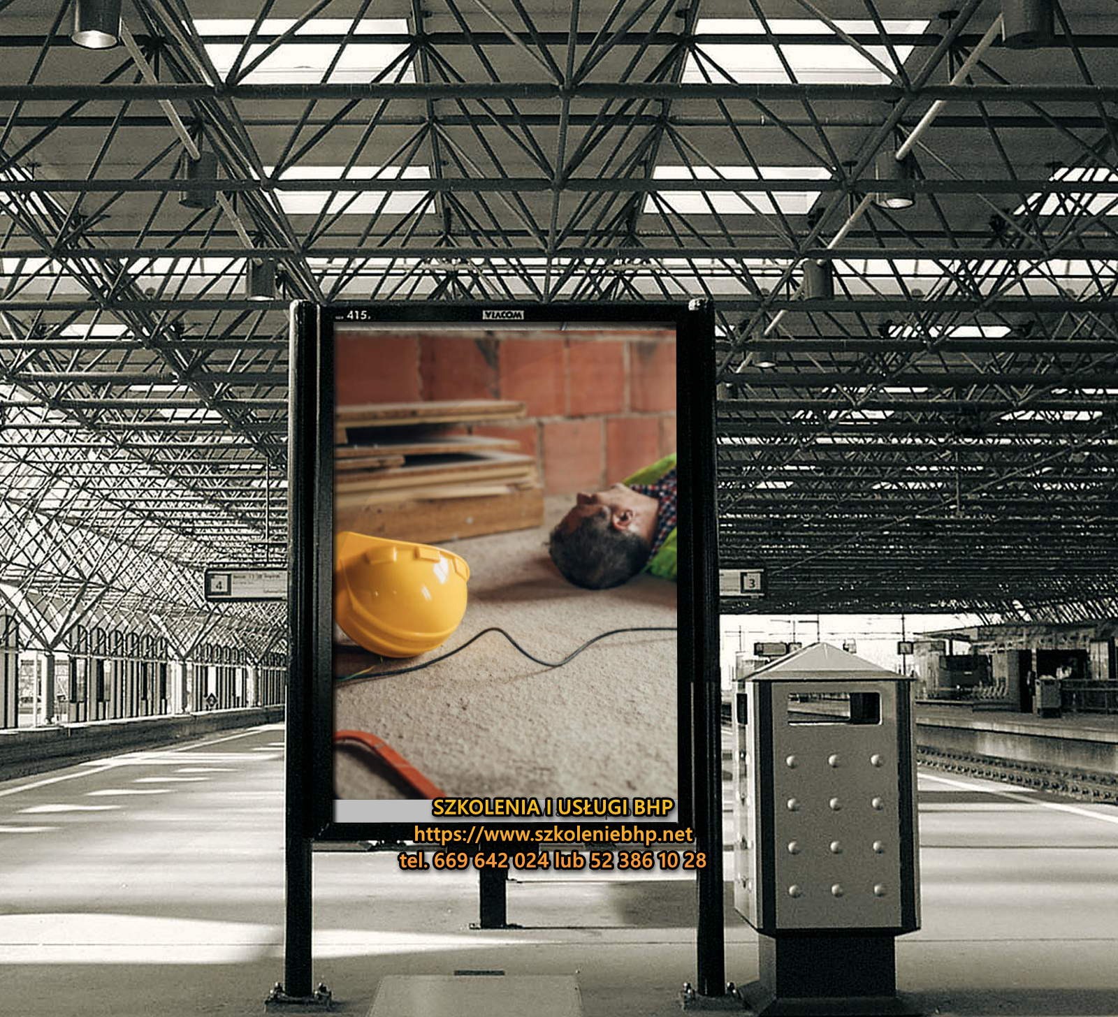 Billboard na dworcu z reklamą przedstawiającą symulację wypadku przy pracy: żółty kask, deski, cegły i leżący mężczyzna.