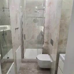 Wykończona łazienka z kabiną prysznicową, podwieszaną toaletą i umywalką w szafce, płytki imitujące marmur na ścianach, widoczne narzędzia i akcesoria do sprzątania na podłodze.