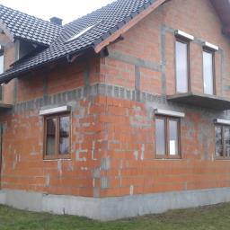 PHU ASTERM Adam Steinhoff - Surowy budynek z czerwonej cegły w trakcie budowy, z zamontowanymi brązowymi oknami z białymi roletami zewnętrznymi, widoczne elementy betonowe fundamentu i nadproży.