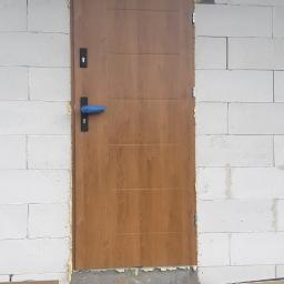 PHU ASTERM Adam Steinhoff - Brązowe drzwi wejściowe z niebieską folią ochronną na klamce, osadzone w surowej ścianie z betonu komórkowego, widoczna izolacja i fundament w trakcie prac wykończeniowych.