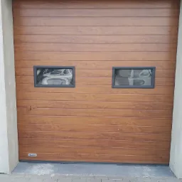 Brązowa segmentowa brama garażowa z dwoma małymi prostokątnymi oknami i poziomym wzorem paneli, widoczny fragment kostki brukowej i logo producenta.