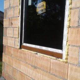 Nowo zamontowane okno z brązowymi ramami i białymi profilami, uszczelnione pianką montażową w murze z cegły ceramicznej.