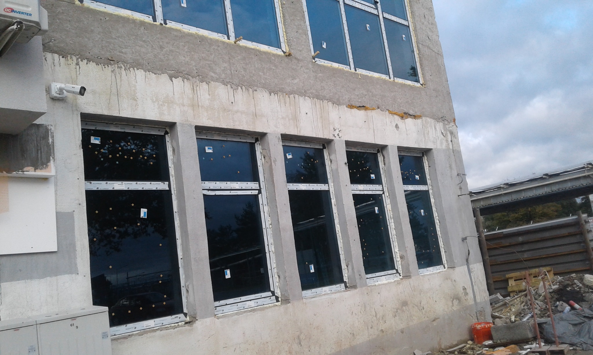 Nowe okna PCV w szarych ramach, zamontowane w surowej elewacji budynku z widocznymi śladami prac budowlanych i ochronną folią na szybach.