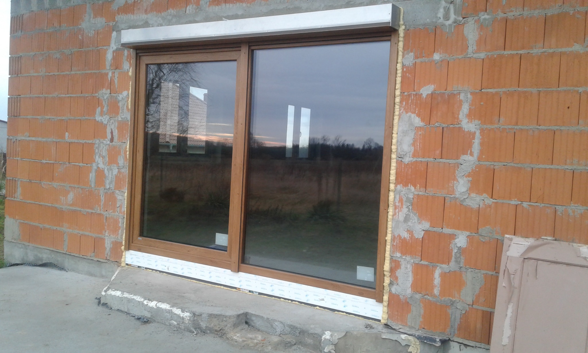 Nowe okno przesuwne, brązowe ramy, pianka montażowa wokół, surowy mur z cegły dziurawki, widok na zewnątrz z odbiciem drzew.