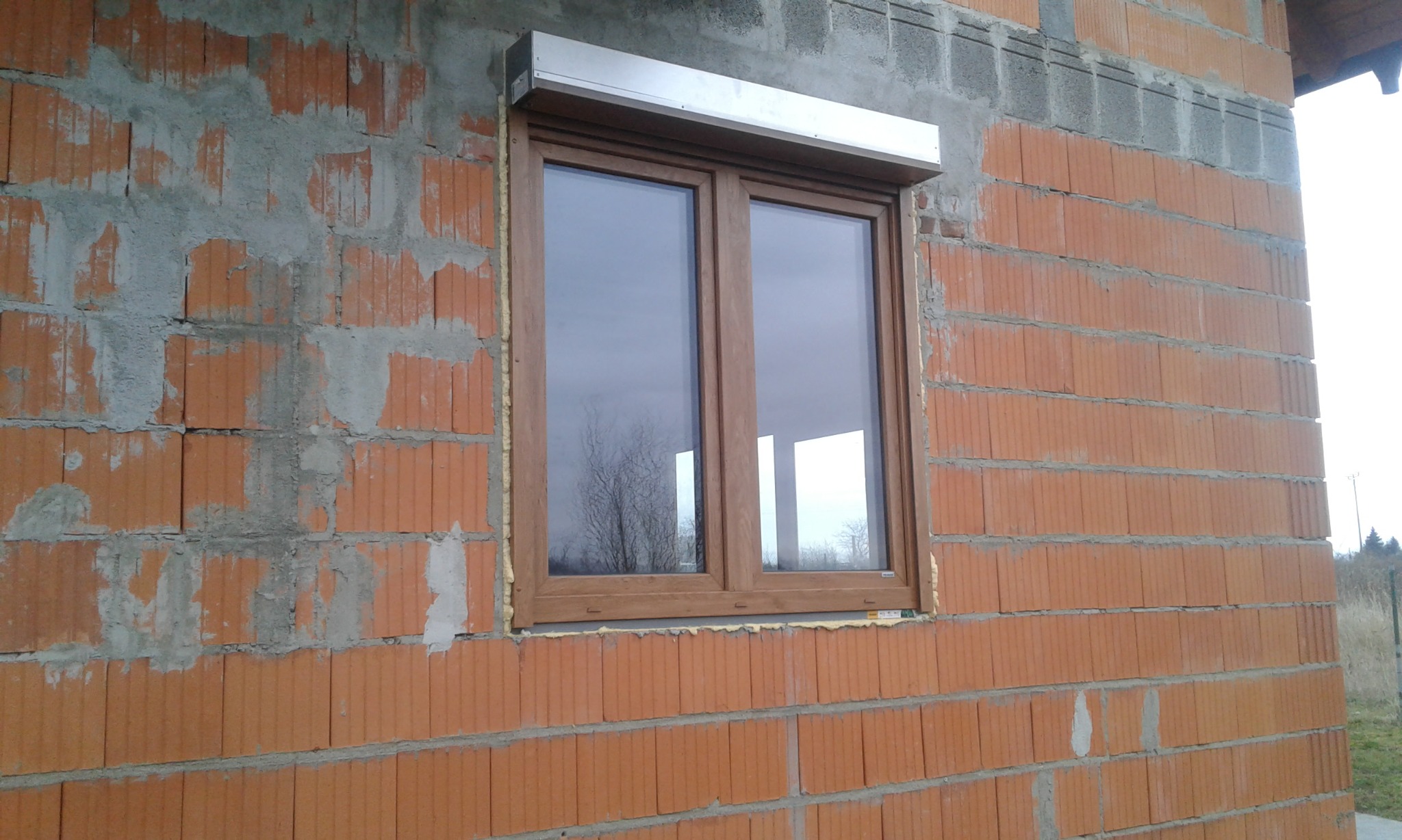 Okna PCV z brązową ramą, zamontowane w surowym murze z czerwonej cegły, z widocznym szarym betonem i pianką montażową, zewnętrzna roleta zwinięta w aluminiowej obudowie.