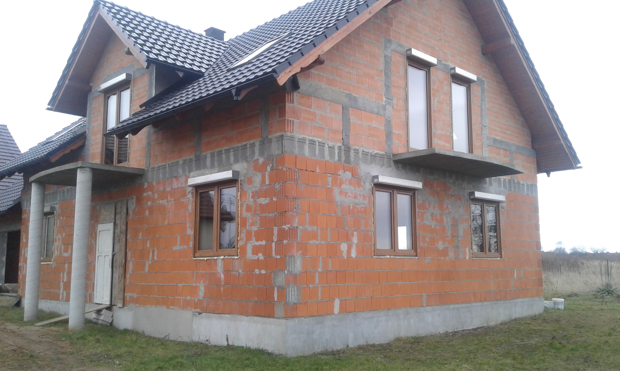 Surowy budynek z czerwonej cegły w trakcie budowy, z zamontowanymi brązowymi oknami z białymi roletami zewnętrznymi, widoczne elementy betonowe fundamentu i nadproży.