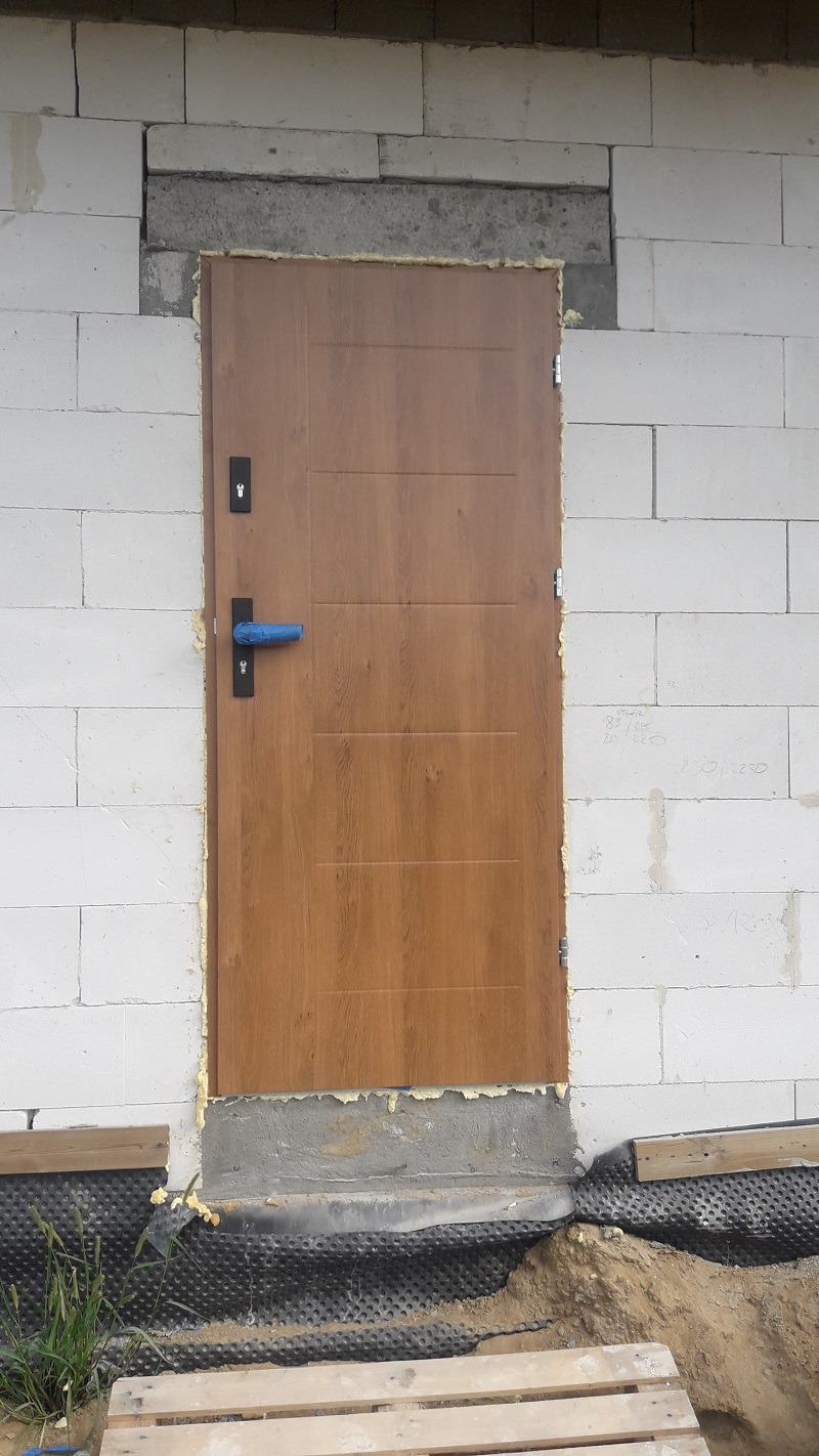 Brązowe drzwi wejściowe z niebieską folią ochronną na klamce, osadzone w surowej ścianie z betonu komórkowego, widoczna izolacja i fundament w trakcie prac wykończeniowych.