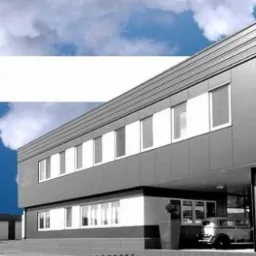 Dwukondygnacyjny budynek biurowy z szarej blachy trapezowej z logotypem HALSTAL i Astron na tle błękitnego nieba z chmurami, widoczna część parkingu z zaparkowanymi samochodami.