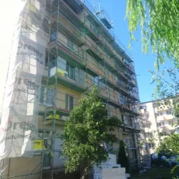 Wielopiętrowy budynek w trakcie prac elewacyjnych, osłonięty rusztowaniami i siatką ochronną z widocznym logo producenta Dreier, z przygotowanymi materiałami izolacyjnymi u podnóża i młodym drzewem...