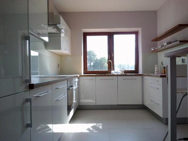 Jasna kuchnia z białymi, lakierowanymi frontami szafek, drewnianym blatem i oknem z widokiem na zieleń, oświetlona naturalnym światłem.