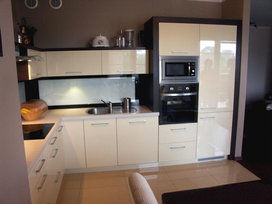 Jasna kuchnia z połyskiem, fronty w kolorze Ral 9010, zabudowa z piekarnikiem i mikrofalówką, ciemne obramowanie szafek górnych i bocznych, płytki podłogowe w ciepłym odcieniu.