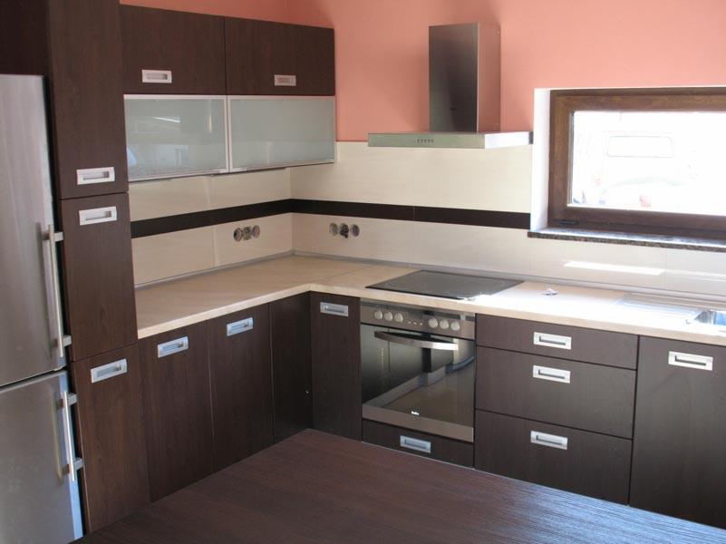 Nowoczesna kuchnia z frontami w kolorze wenge, jasnymi blatami i płytkami, oraz srebrnymi uchwytami. Widoczny sprzęt AGD: lodówka, piekarnik i płyta indukcyjna. Nad blatem roboczym okap ze stali...