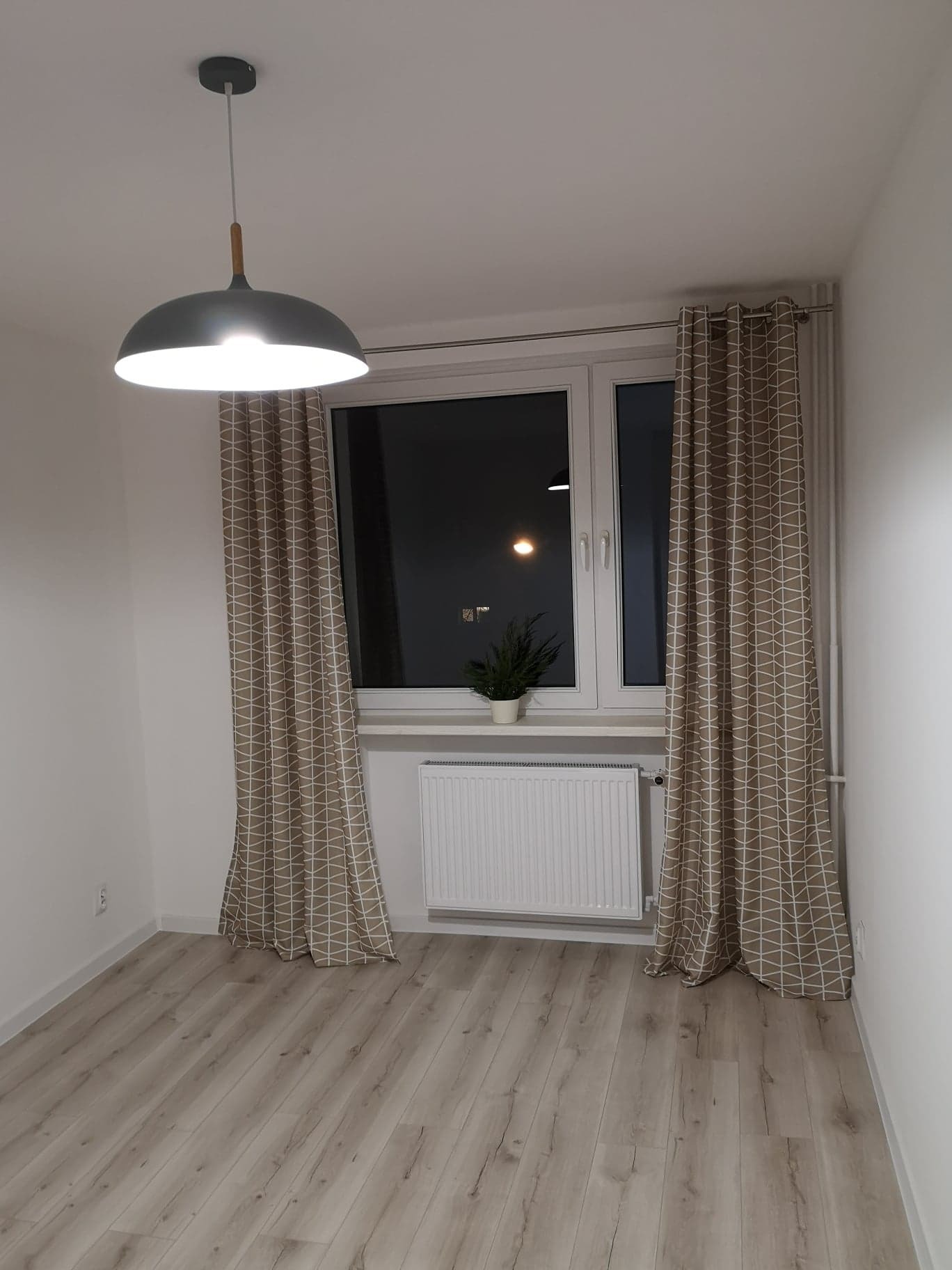 Pusty pokój z panelami podłogowymi w odcieniach szarości, grzejnikiem pod oknem z rośliną w doniczce, szarą lampą wiszącą i zasłonami w geometryczny wzór.