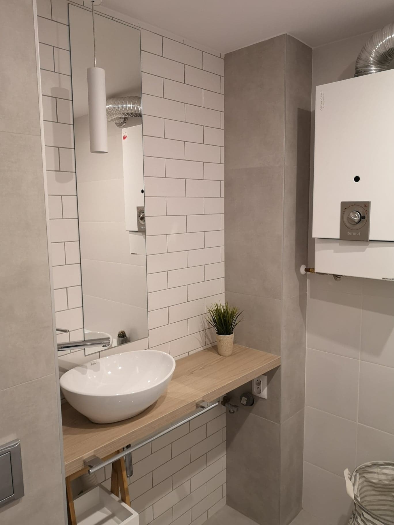 Nowoczesna łazienka z białą umywalką nablatową na drewnianym blacie, lustro w tle odbija biały grzejnik łazienkowy, ściany wyłożone białymi i szarymi płytkami.