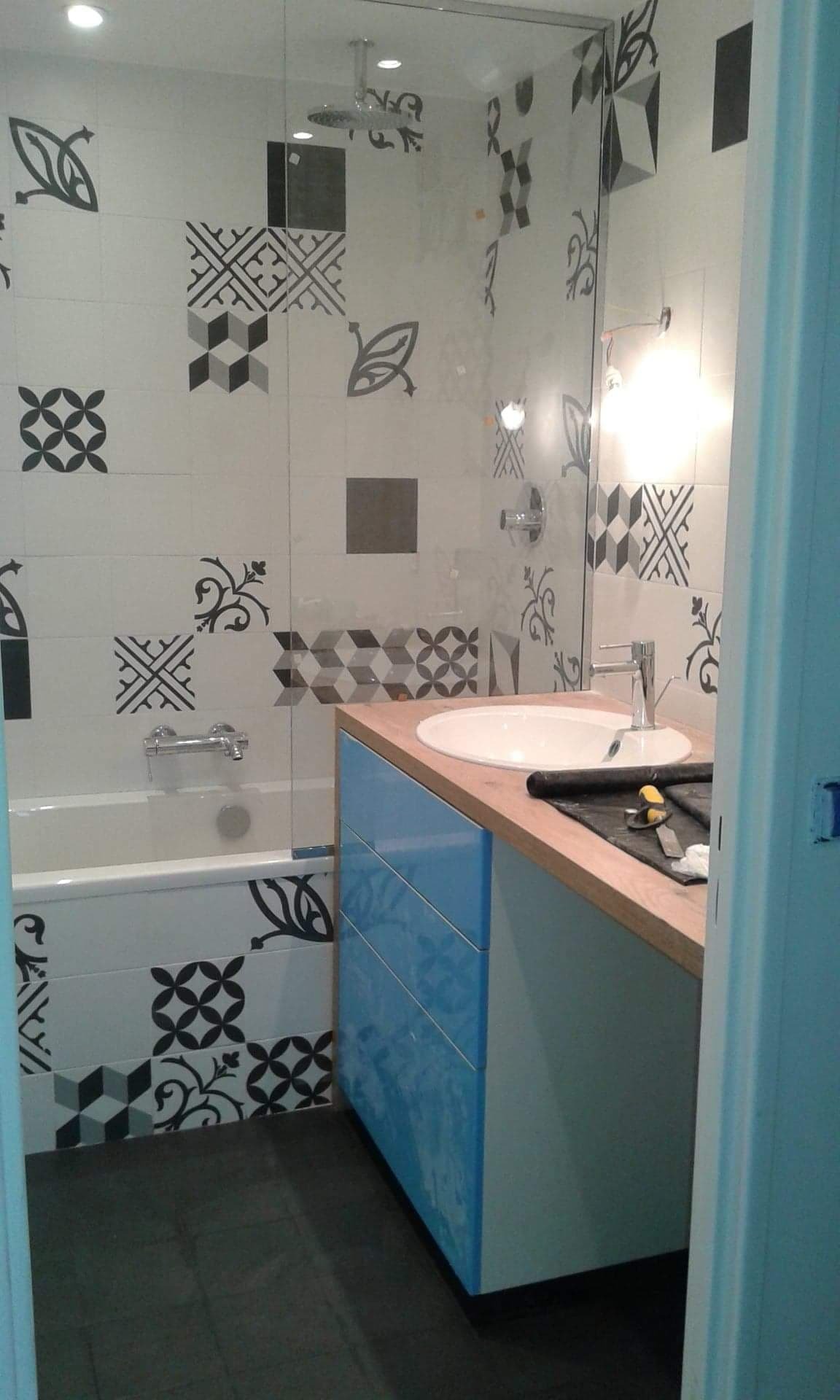 Wykończona łazienka z wanną obudowaną płytkami w geometryczne i roślinne wzory, kabina prysznicowa ze szklaną ścianką i umywalką z szafką w odcieniach błękitu i drewna. Widoczne narzędzia na blacie.