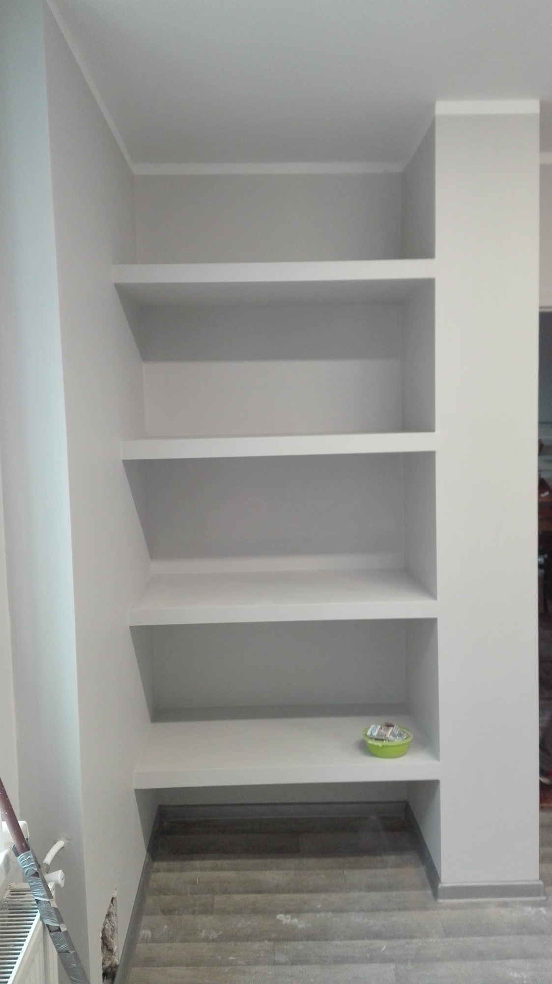 Wnęka z czterema półkami pomalowana na biało, na dolnej półce zielona miseczka z drobiazgami, szara podłoga imitująca drewno, widoczne ślady po remoncie na ścianie przy grzejniku.