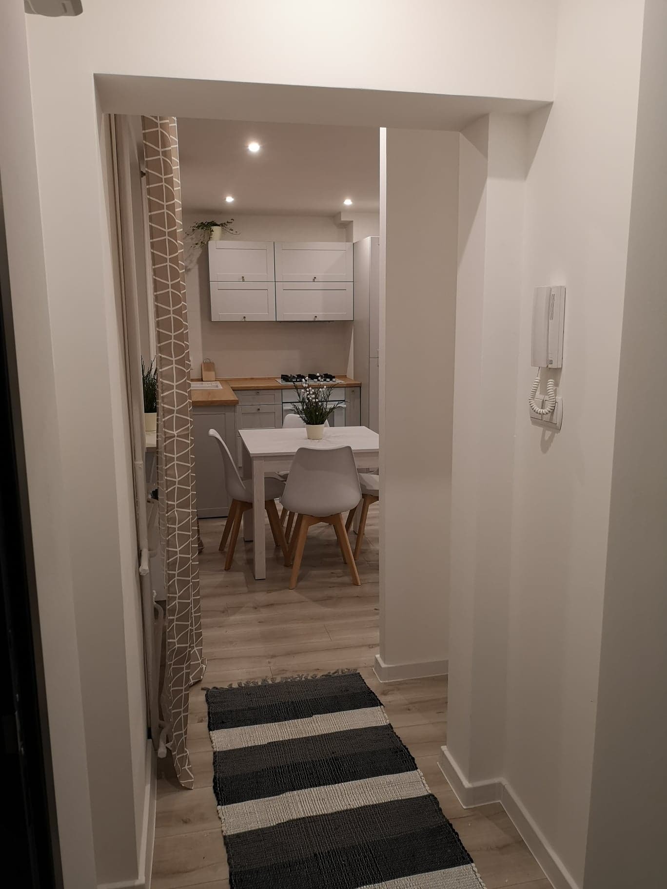Widok na jasną kuchnię z jadalnią z perspektywy przedpokoju; widoczne białe ściany, parkiet, stół z krzesłami i dywan w paski przy wejściu.