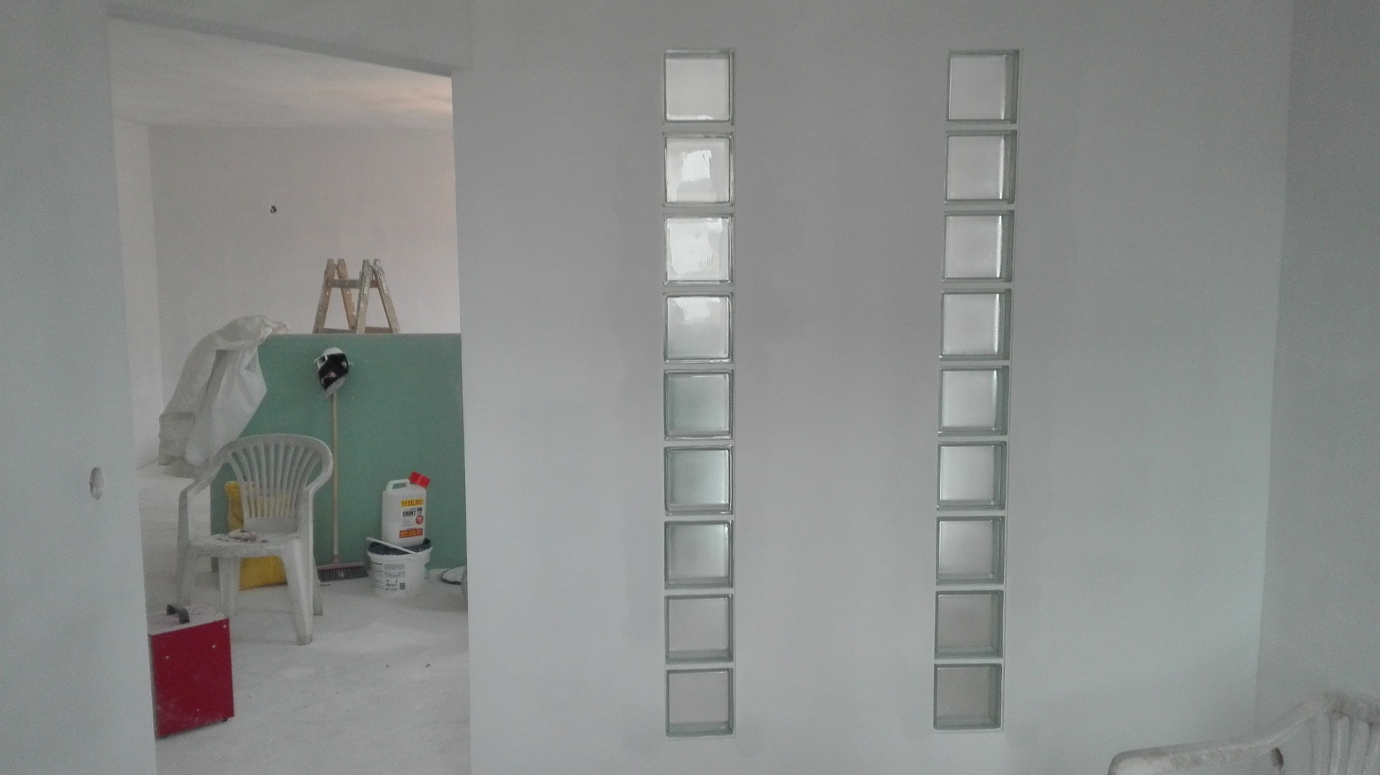 Biała ściana z dwoma pionowymi rzędami szklanych pustaków, widok na remontowane pomieszczenie z drabiną i materiałami budowlanymi w tle.