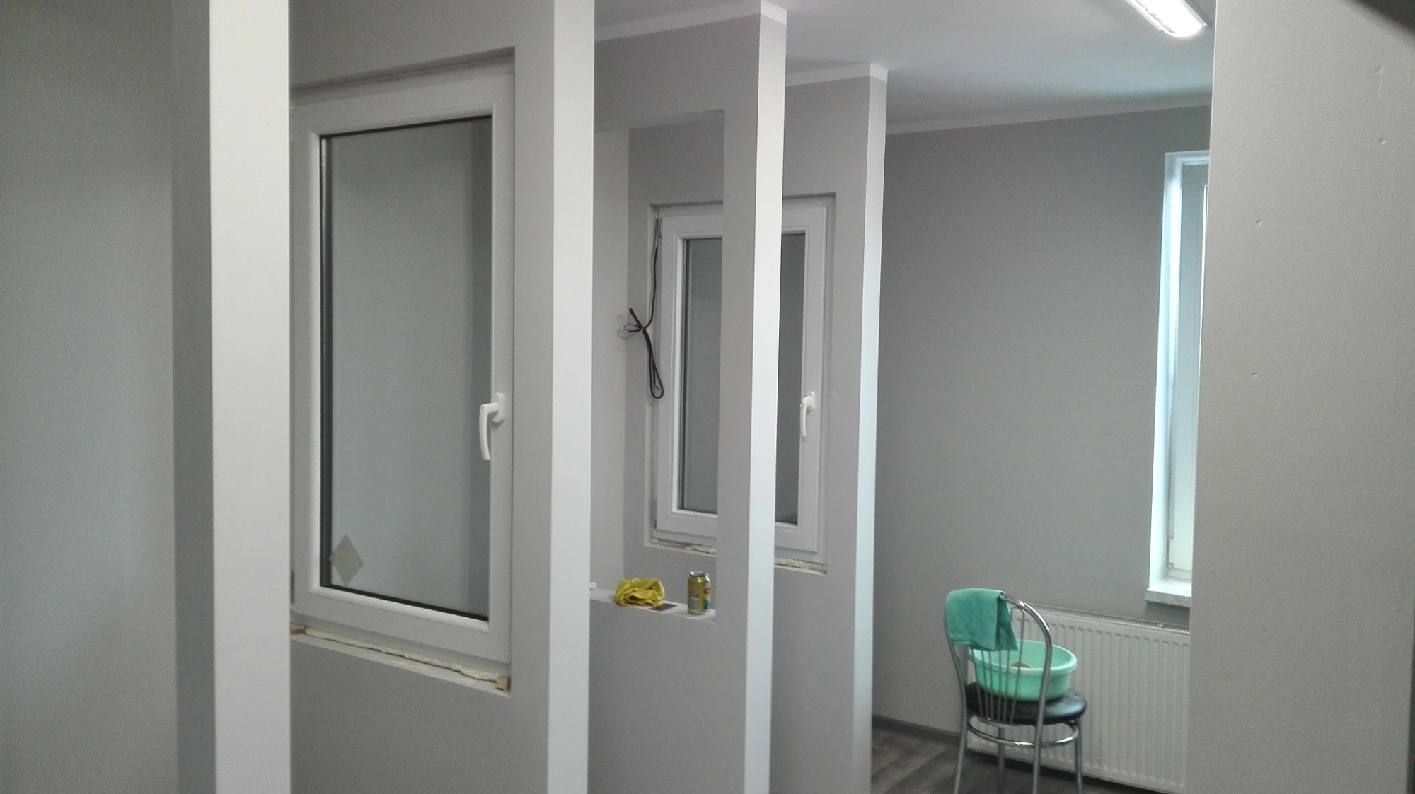 Szare wnętrze z trzema oknami oddzielonymi białymi kolumnami, widoczne ślady uszczelniania okien, na półce pod środkowym oknem leżą żółta szmatka i puszka, obok krzesło z zielonym ręcznikiem.