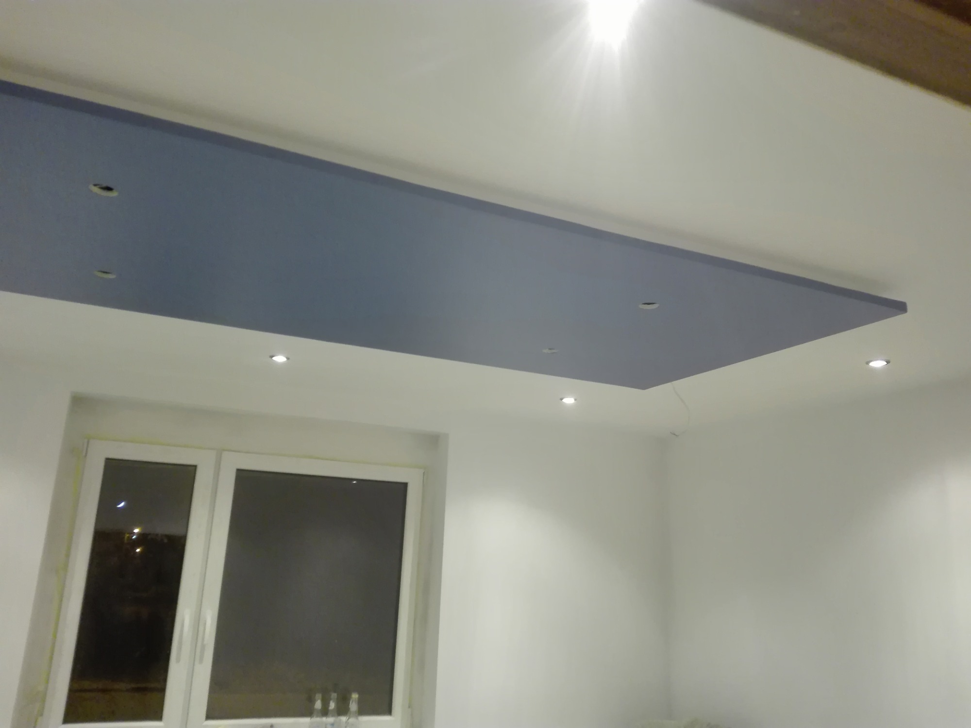 Niebieski, prostokątny element sufitu podwieszanego z otworami na oświetlenie punktowe, zamontowany w białym pomieszczeniu z oknem.