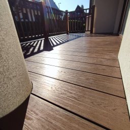 DK BUILDING - Nowy taras kompozytowy w słoneczny dzień. Widoczne deski tarasowe, balustrada i cień. Na pierwszym planie duża donica. Perspektywa z dołu.