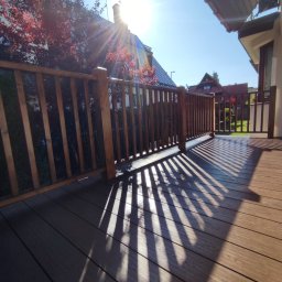 DK BUILDING - Nowoczesny taras kompozytowy z drewnianą balustradą w Dobra, oświetlony promieniami słońca tworzącymi ciekawe cienie. Widok na dom i drzewa w tle.