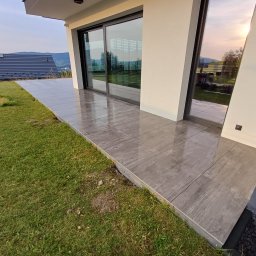 DK BUILDING - Nowoczesny taras z szarych płytek imitujących drewno, przylegający do budynku z dużymi oknami przesuwnymi, otoczony zielonym trawnikiem i widokiem na wzgórza.