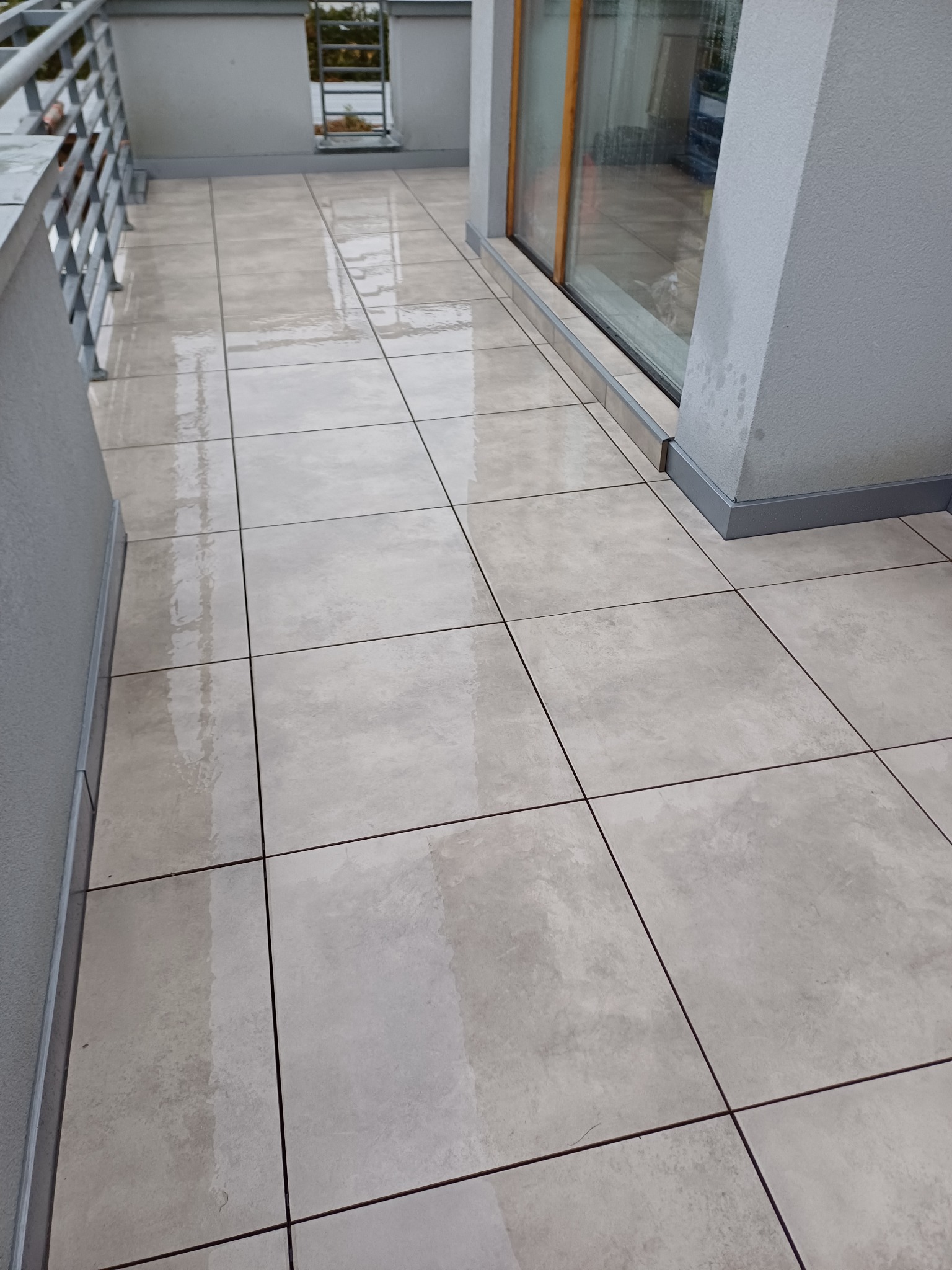 Jasne, kwadratowe płytki na balkonie, ułożone równo, z widocznymi fugami. Balustrada i szare ściany w tle. Widoczne ślady wilgoci na powierzchni płytek.