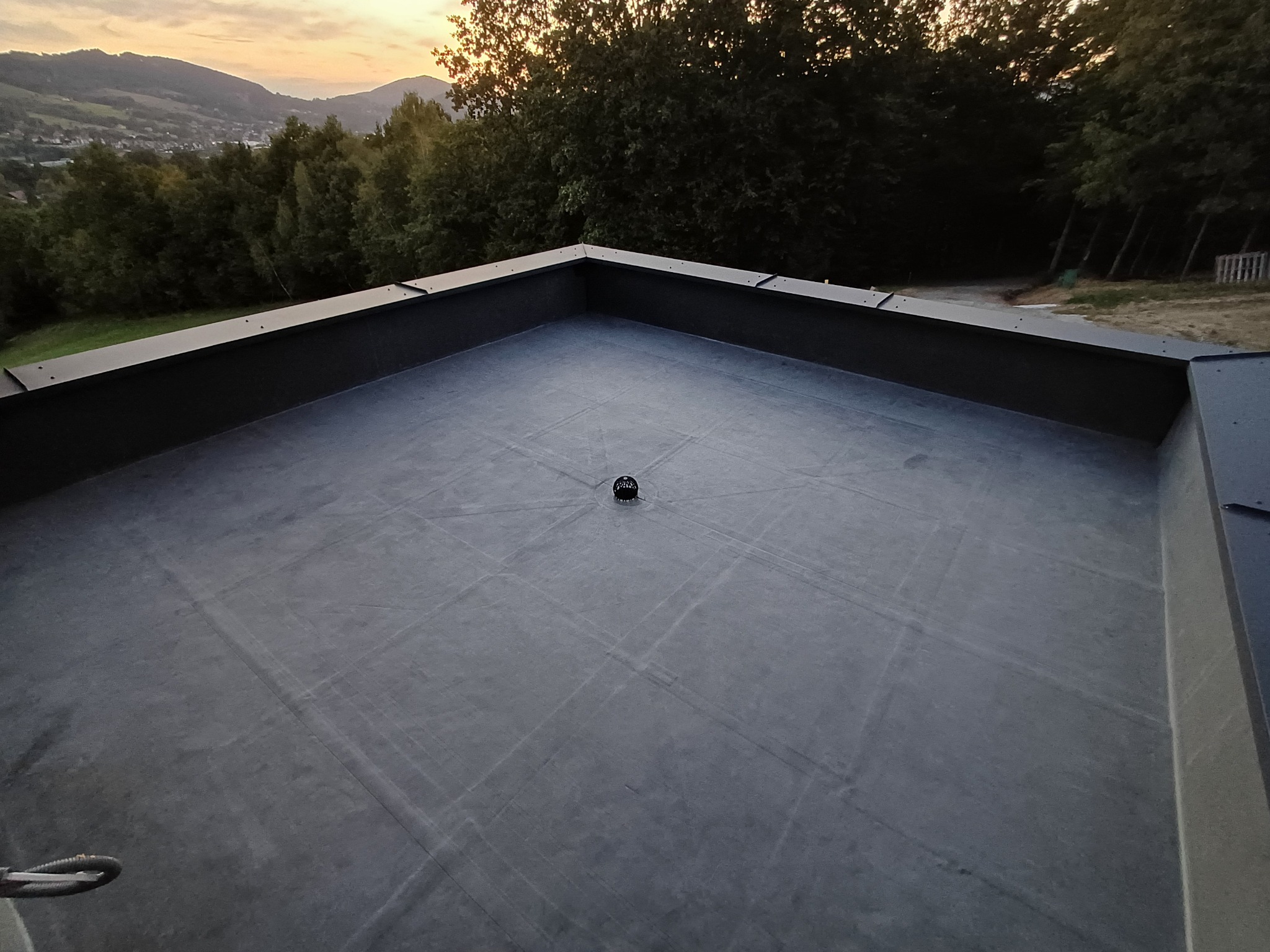 Szary, płaski dach z odpływem, otoczony ciemną obróbką blacharską. W tle drzewa i pagórki w wieczornym świetle.