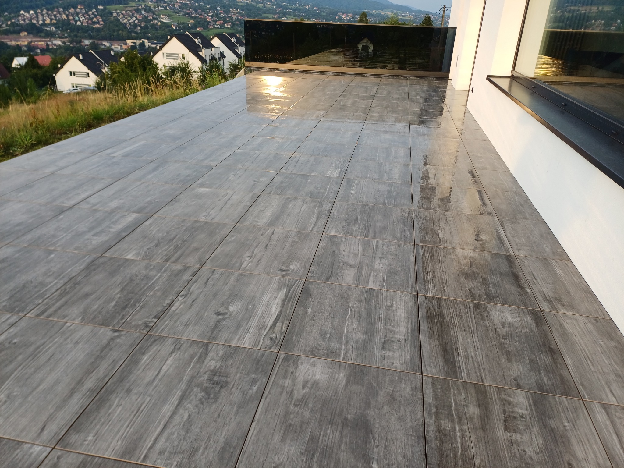 Nowoczesny taras wyłożony szarymi płytkami imitującymi drewno, z widokiem na miasto i góry w tle. Minimalistyczna balustrada ze szkła dodaje elegancji.