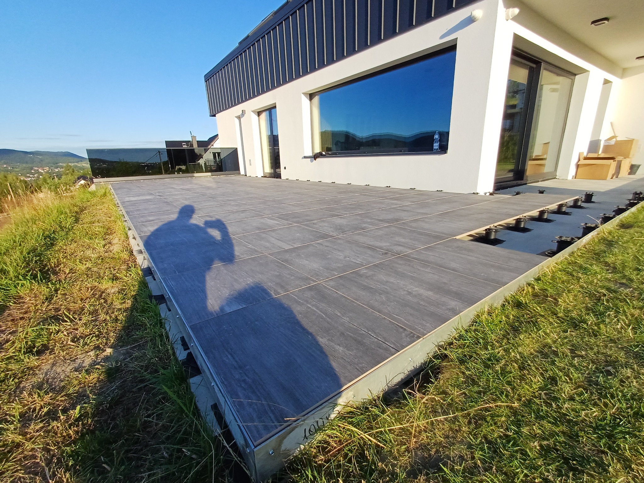 Nowoczesny taras z szarych płyt na regulowanych wspornikach, z widokiem na góry i dom. Czyste linie i minimalistyczny design.