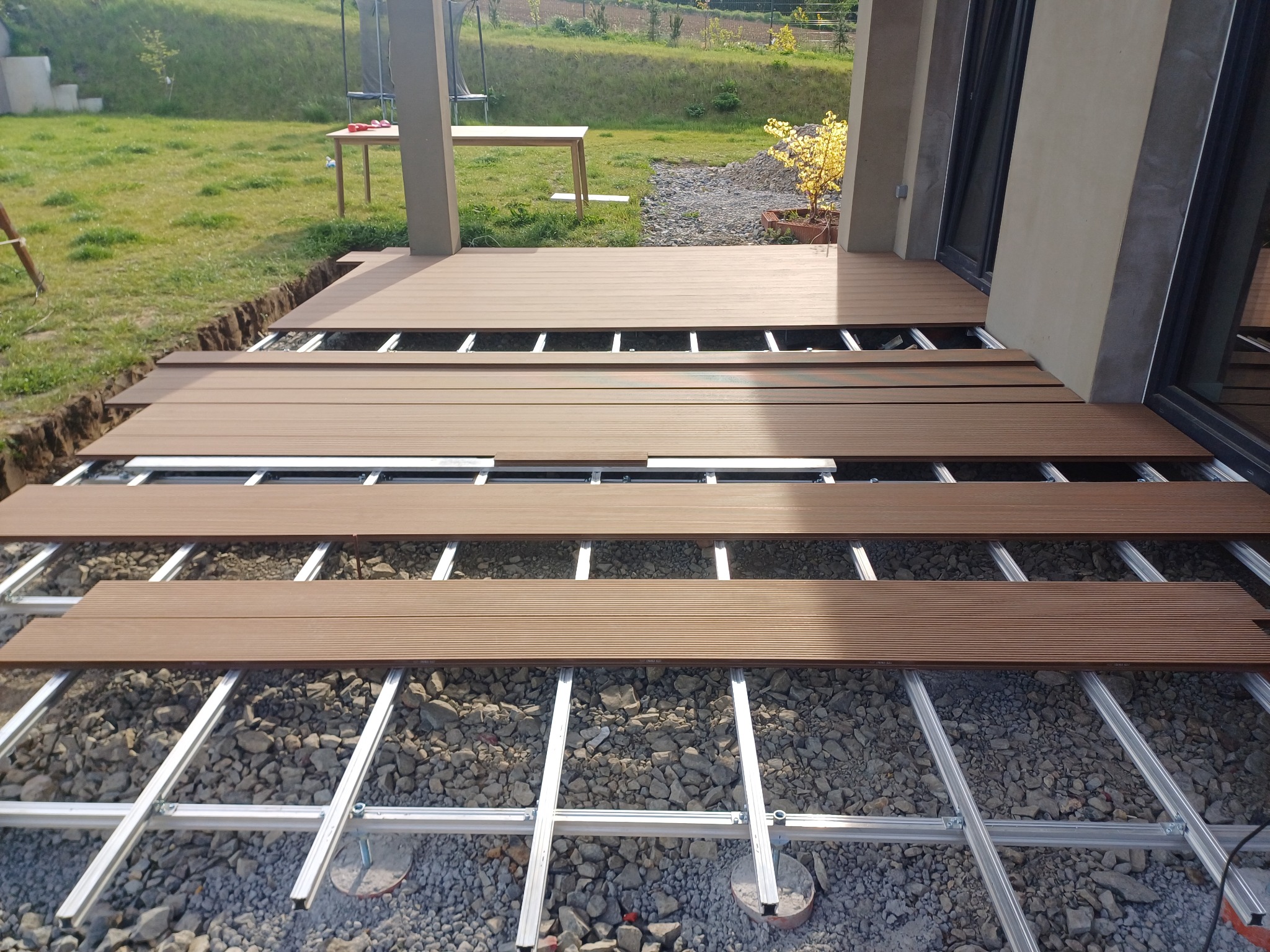 Montaż tarasu kompozytowego: widoczne legary aluminiowe i deski w kolorze drewna na podłożu z kamieni. Część tarasu już ułożona, reszta w trakcie montażu.