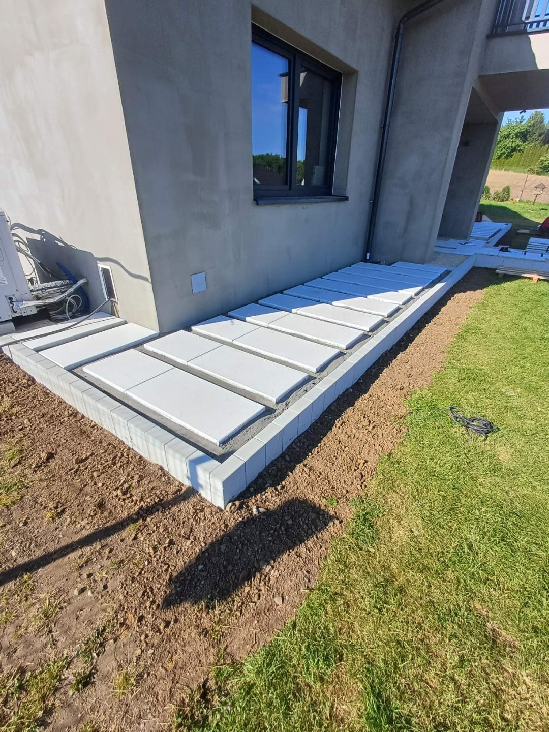 Montaż tarasu z betonowych płyt na zewnątrz domu. Widoczne równe ułożenie płyt i obrzeża. Częściowo wykończony taras na trawniku, etap prac.