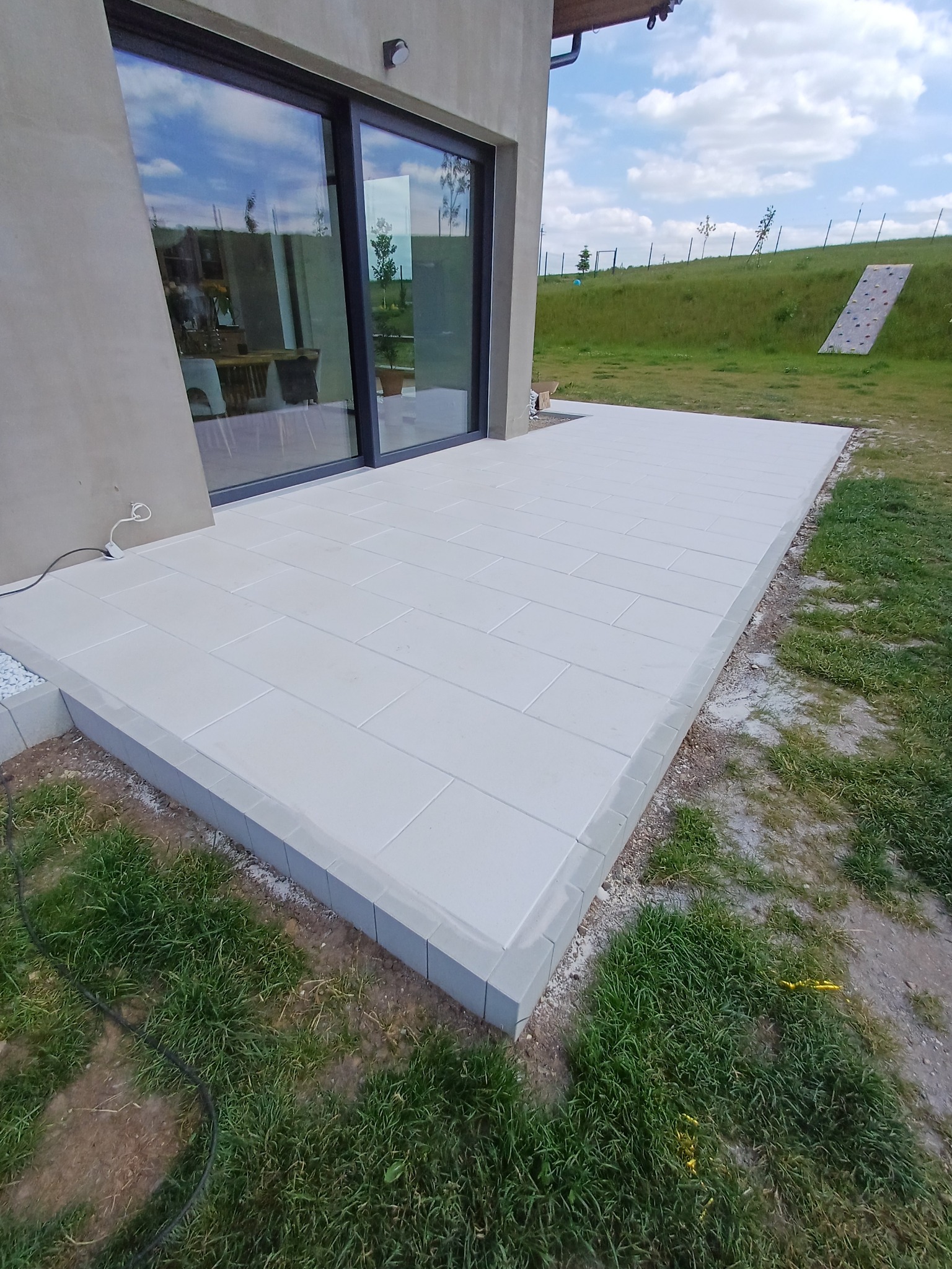 Jasny taras z betonowych płyt, z obrzeżem z szarych kostek, przylegający do domu z dużym oknem przesuwnym i zielonym trawnikiem w tle.