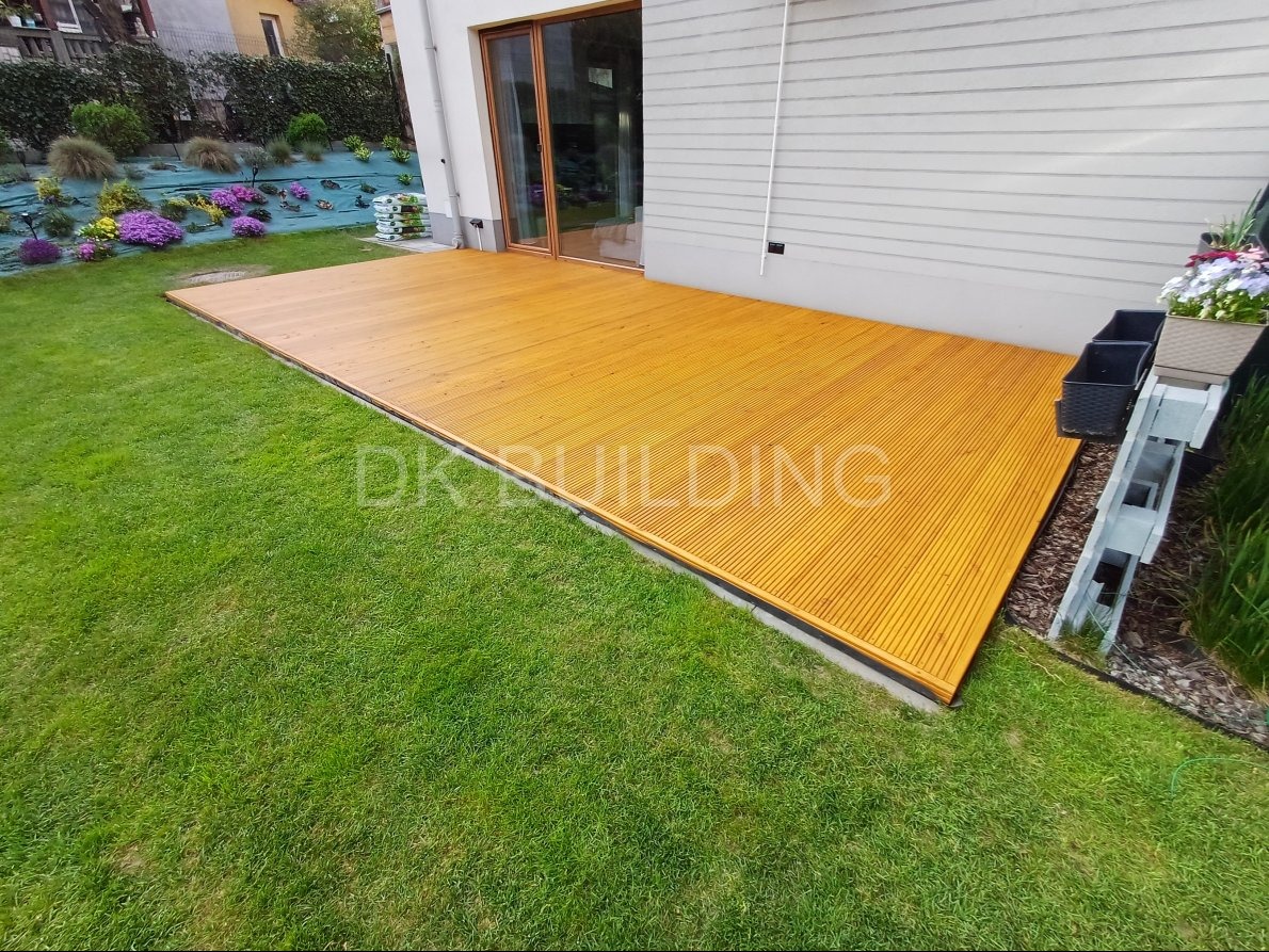 Nowoczesny taras z drewna egzotycznego przylegający do domu, otoczony zielonym trawnikiem i rabatami kwiatowymi. Widoczne donice z kwiatami.