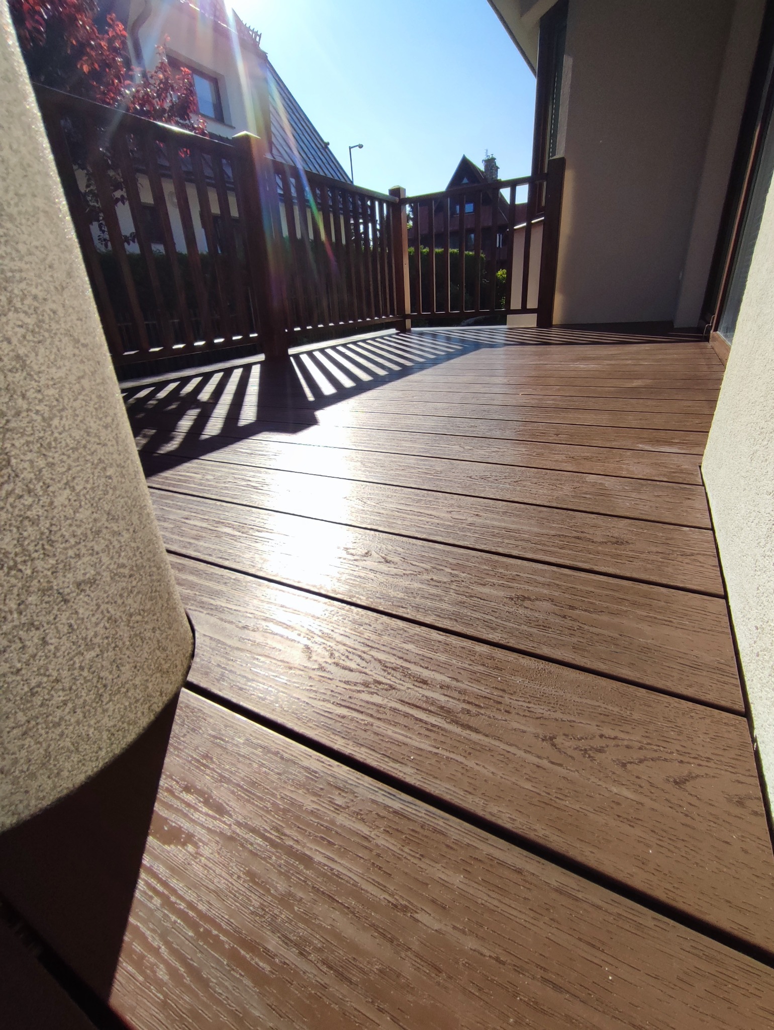 Nowy taras kompozytowy w słoneczny dzień. Widoczne deski tarasowe, balustrada i cień. Na pierwszym planie duża donica. Perspektywa z dołu.