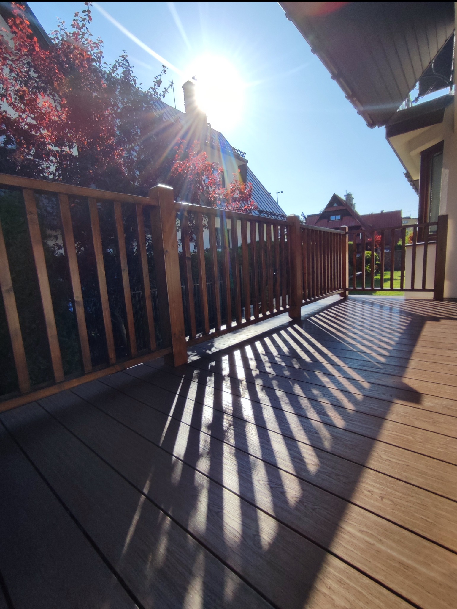 Nowoczesny taras kompozytowy z drewnianą balustradą w Dobra, oświetlony promieniami słońca tworzącymi ciekawe cienie. Widok na dom i drzewa w tle.