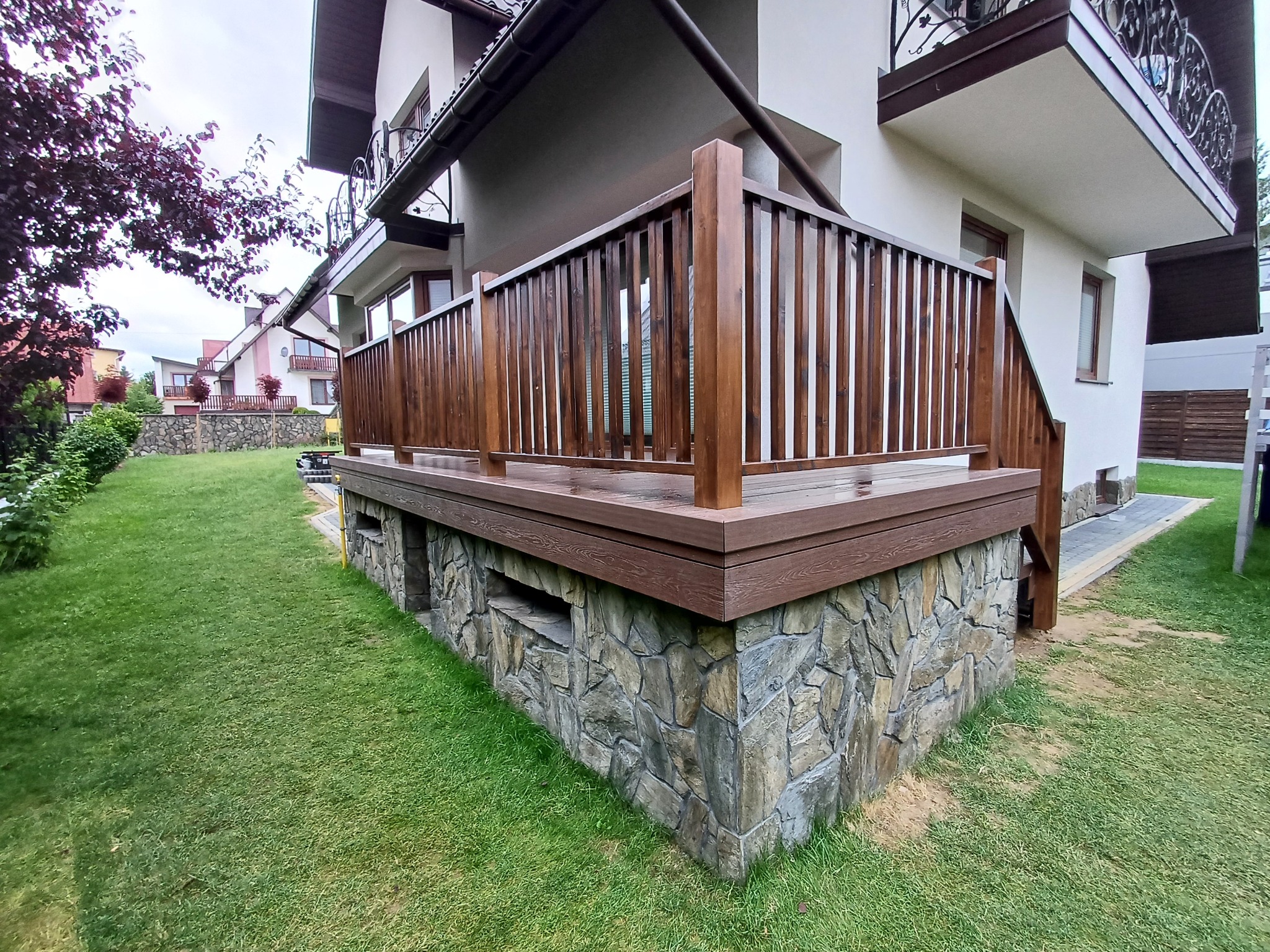 Nowoczesny taras z brązową deską kompozytową i kamienną podmurówką, widok z poziomu trawnika, na tle domu z balkonem.
