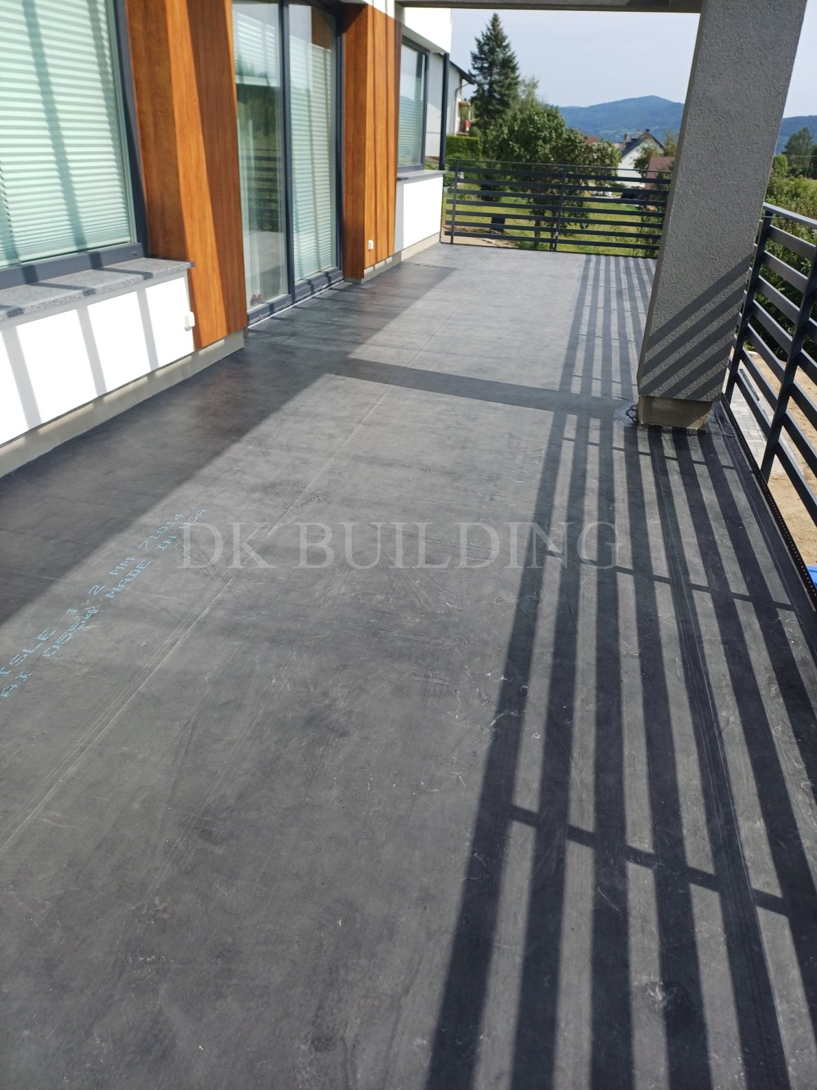 Nowoczesny taras betonowy z widokiem na góry, czarna, gładka powierzchnia, cień barierki, jasne otoczenie, widoczne napisy techniczne na podłożu.