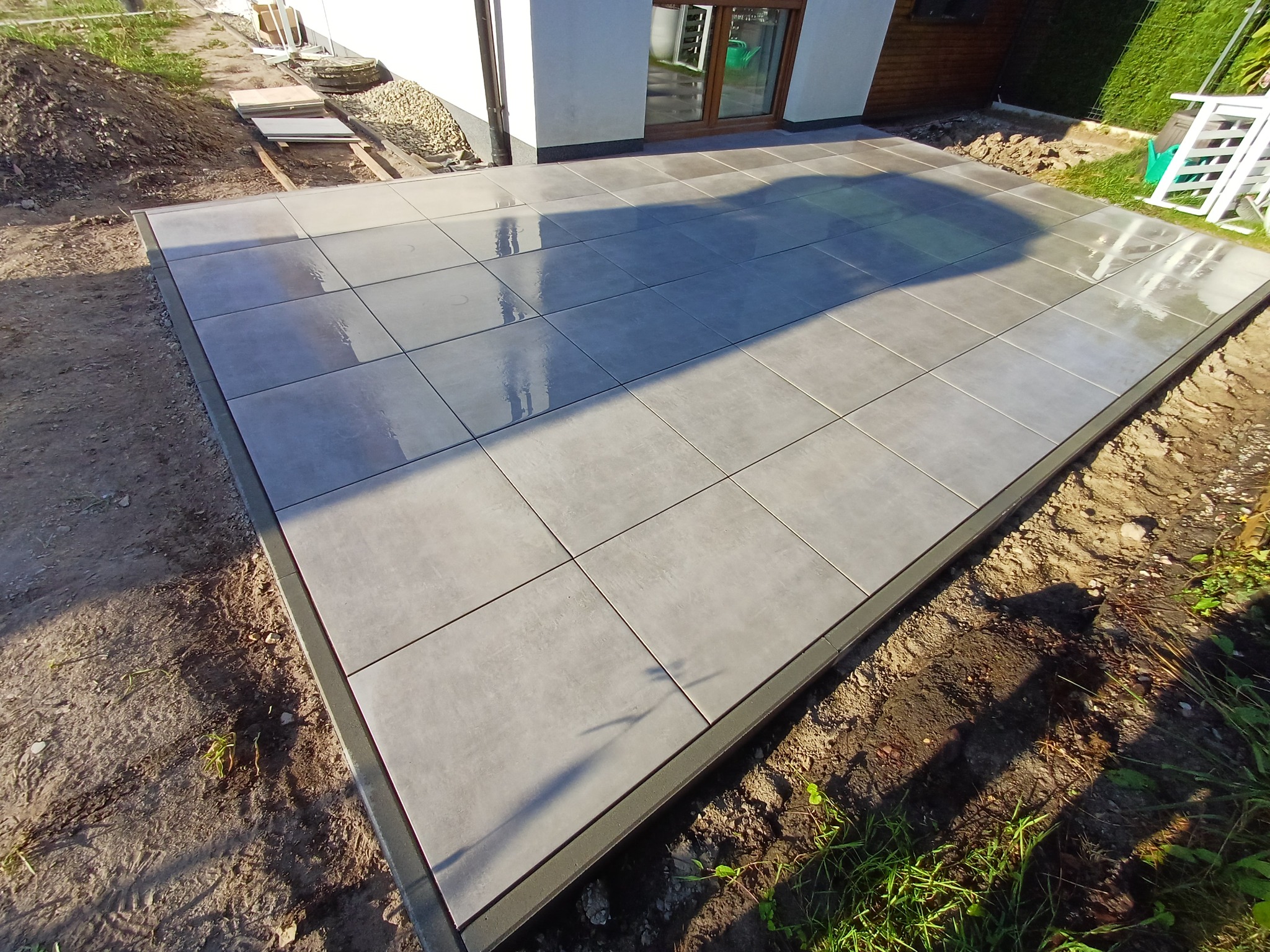 Nowoczesny taras z szarych płyt na gruncie, obramowany ciemną listwą. Widoczne przygotowania do wykończenia terenu wokół tarasu. Świeżo ułożona nawierzchnia.