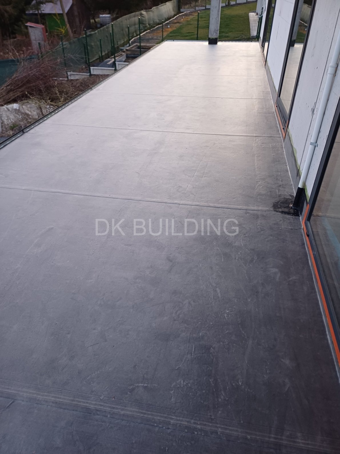 Gładka, szara powierzchnia tarasu betonowego z widocznymi liniami podziału. W tle zieleń i elementy konstrukcyjne budynku z oknami.