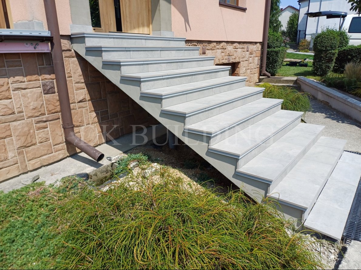 Zewnętrzne schody wejściowe wyłożone jasnoszarą płytką z delikatnym obramowaniem, prowadzące do drzwi, z widoczną rynną i zielenią w tle.