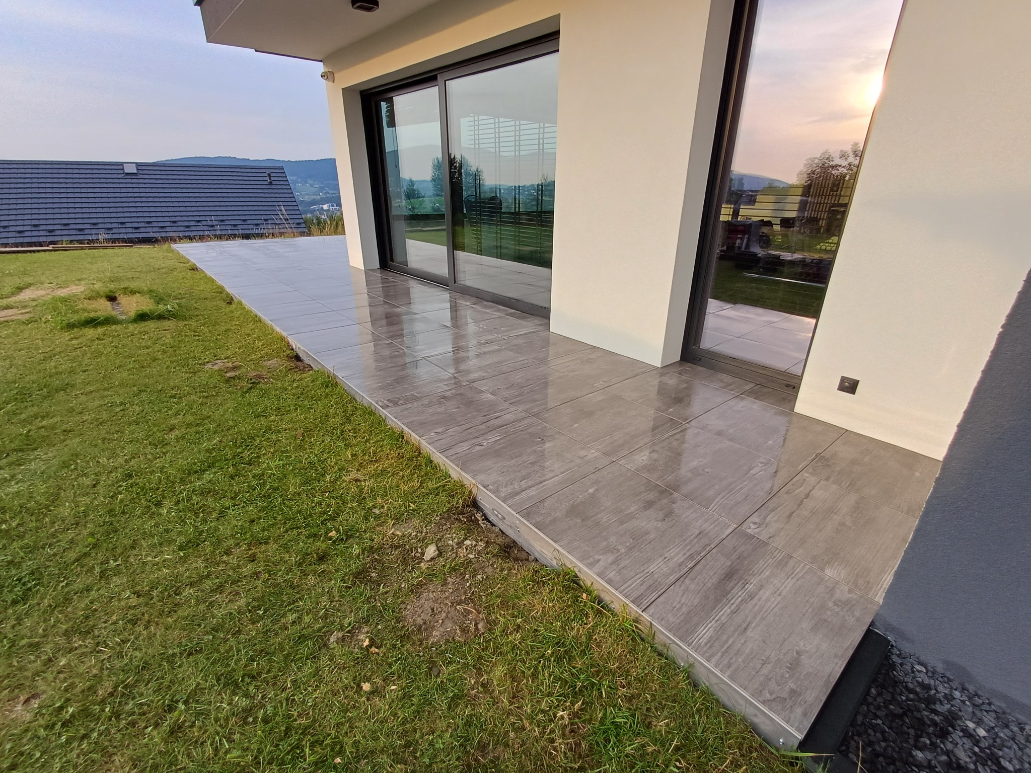 Nowoczesny taras z szarych płytek imitujących drewno, przylegający do budynku z dużymi oknami przesuwnymi, otoczony zielonym trawnikiem i widokiem na wzgórza.
