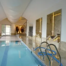 Kryty basen z marmurowym wykończeniem ścian i podłogi. Oświetlone nisze ścienne, spiralne schody w tle. Częściowo zakryty basen z błękitną wodą.