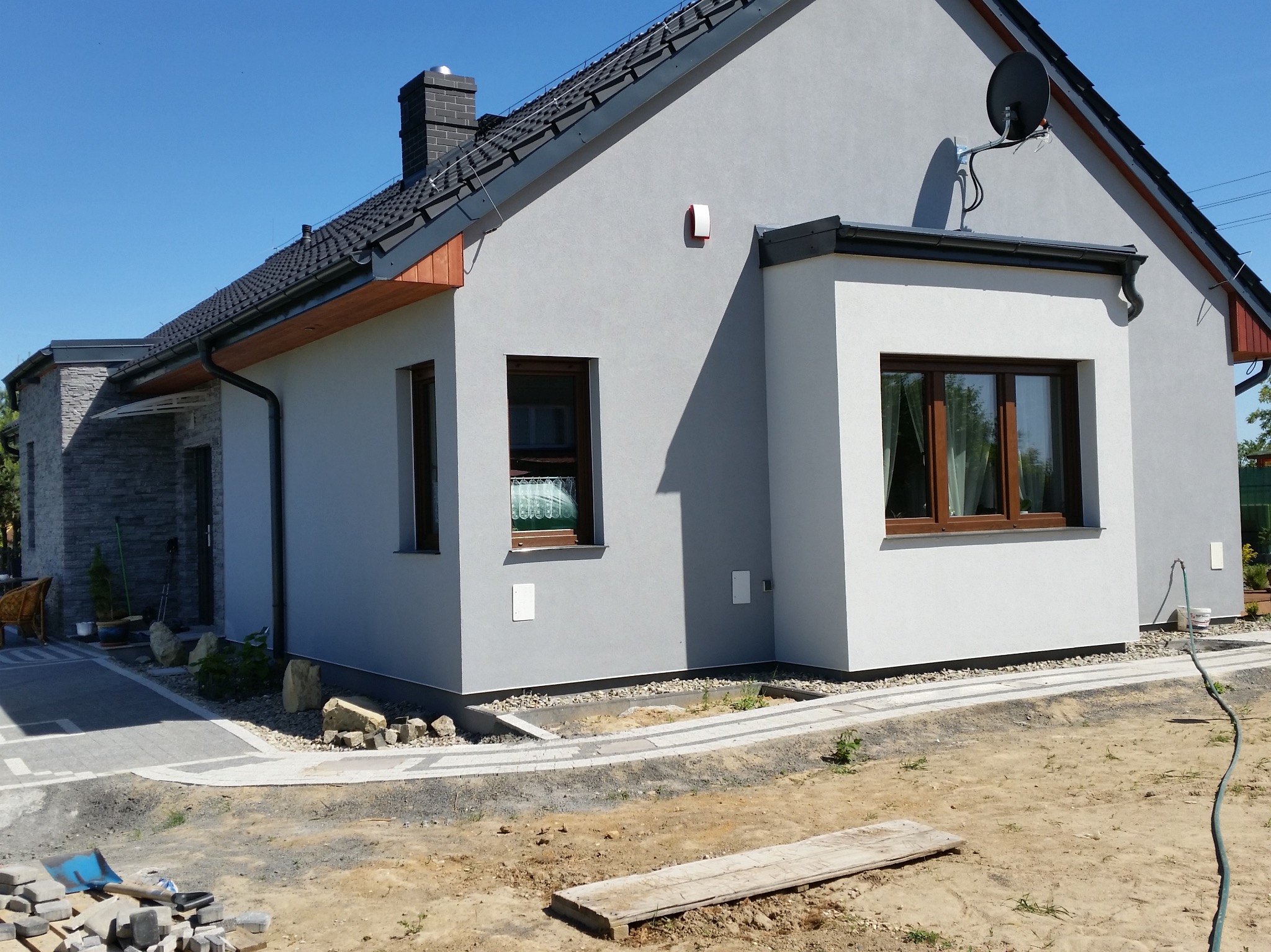 Nowoczesny dom jednorodzinny z szarą elewacją, czarnym dachem i kamiennym wykończeniem fragmentu ściany. Widoczna kostka brukowa i teren w trakcie prac wykończeniowych.