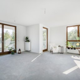Dominor Nieruchomości - Jasne, przestronne wnętrze apartamentu z dużymi oknami i betonową podłogą. Widok na zieleń za oknem, minimalistyczny wystrój z roślinami i fotel z narzutą.