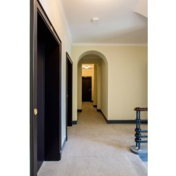 Dominor Nieruchomości - Korytarz w kamienicy z ciemnymi drzwiami i łukowym przejściem. Widoczne schody z metalową balustradą. Stonowana kolorystyka ścian i podłogi.